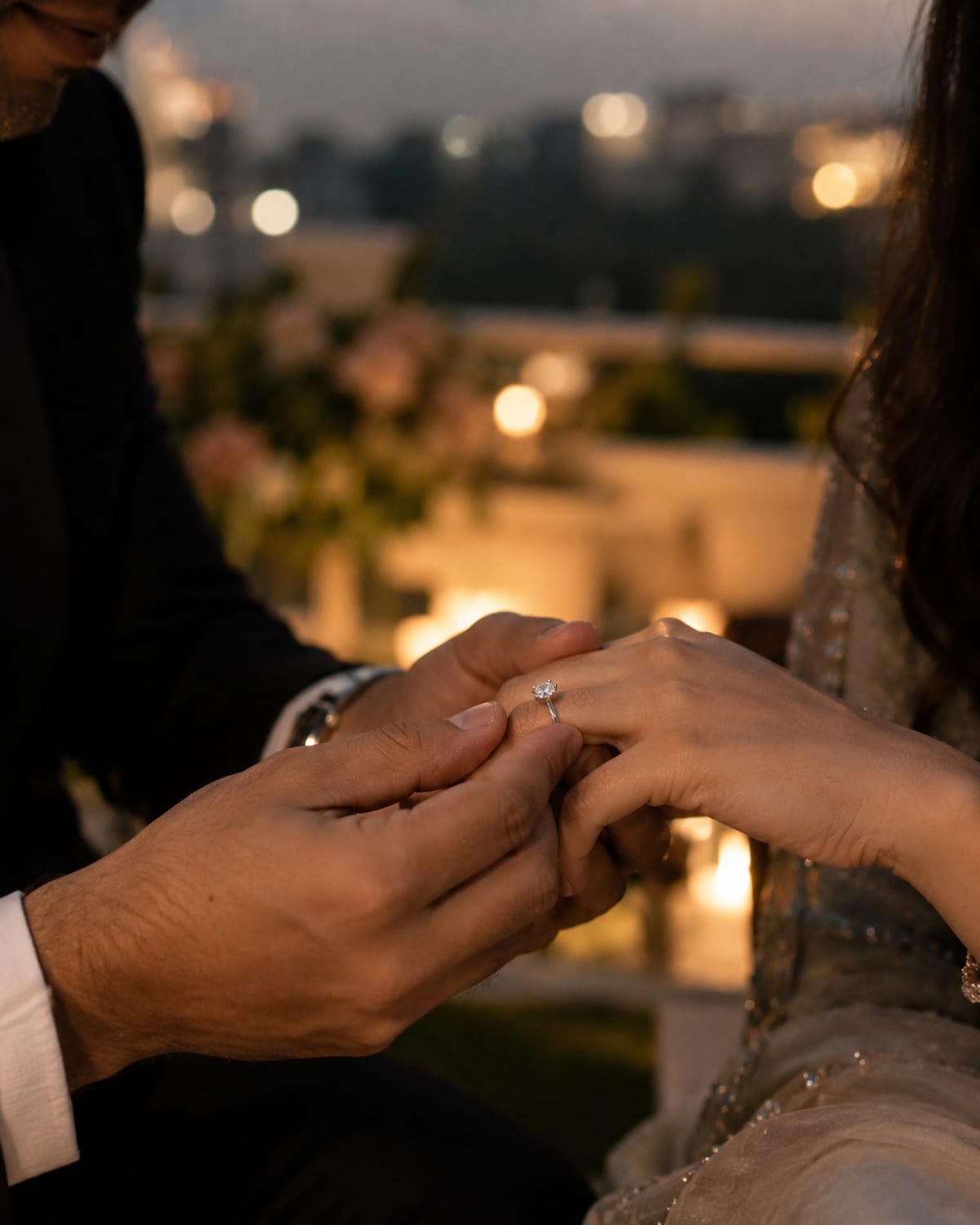 Vikram slipping a diamond engagement ring onto Ananya's finger at dusk, city lights blurred behind them